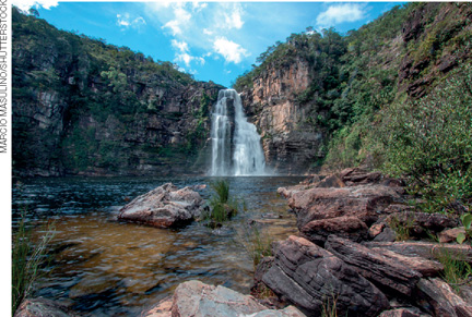 IMAGEM: uma grande cachoeira cai de um paredão de rochas com vegetação e, embaixo, forma um rio, cercado por pedras e mais vegetação. FIM DA IMAGEM.