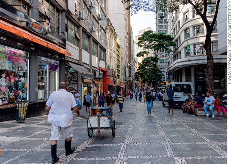 IMAGEM: uma rua larga com calçamento de pedra, cercada por prédios e lojas e por onde circulam pessoas e vendedores ambulantes. FIM DA IMAGEM.