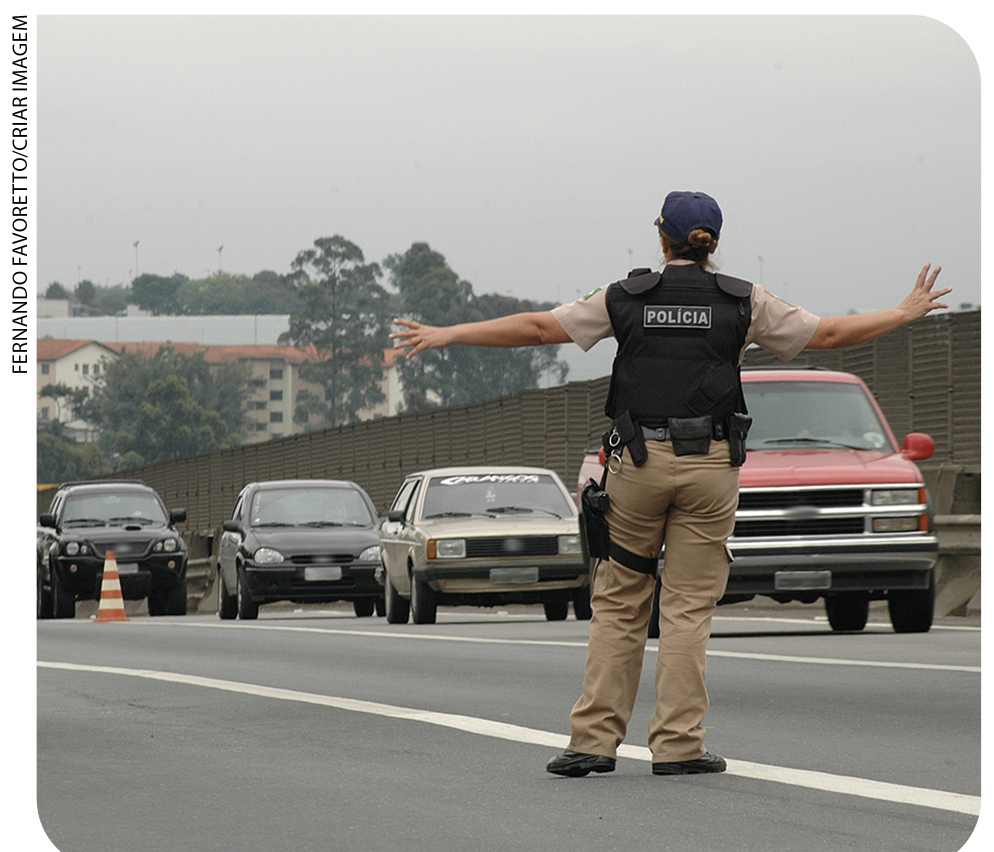 IMAGEM: uma mulher com colete da polícia e boné controla o trânsito. FIM DA IMAGEM.