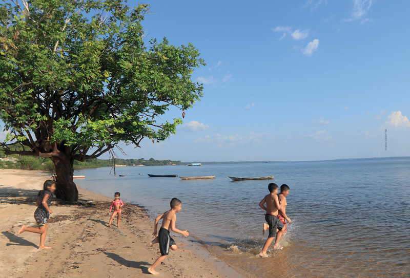 IMAGEM: crianças molhadas brincam à beira do rio, que está cercado por vegetação e onde há vários barcos. FIM DA IMAGEM.