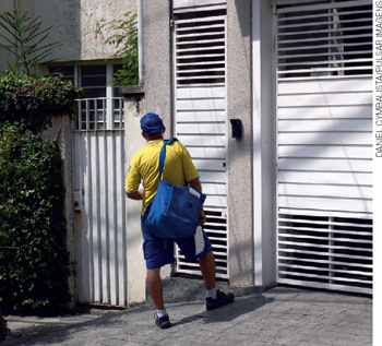 IMAGEM: um homem de uniforme amarelo e azul e com uma sacola de cartas no ombro está na frente de uma casa. FIM DA IMAGEM.