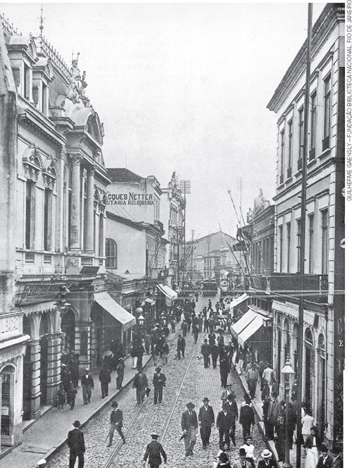 IMAGEM: fotografia antiga, em preto e branco, de uma rua comercial, com grandes fachadas e calçamento de paralelepípedo, pela qual caminham homens com chapéus e vestes da época. FIM DA IMAGEM.