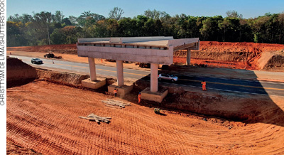 IMAGEM: construção de um viaduto sobre uma rodovia. ele é sustentado por grandes pilastras e tem terra dos seus dois lados, às margens da rodovia. FIM DA IMAGEM.