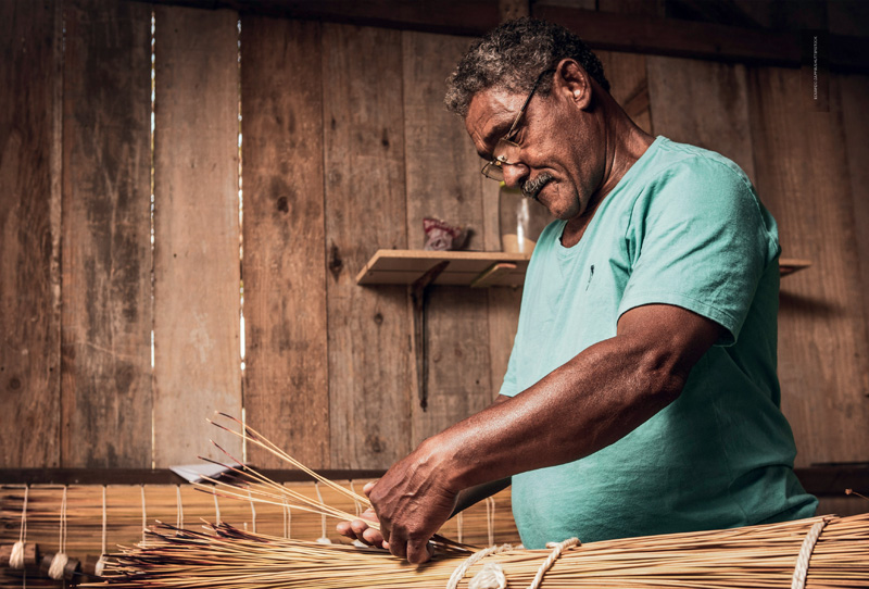 IMAGEM: um homem produz uma esteira alinhando e amarrando fibras de palha. FIM DA IMAGEM.