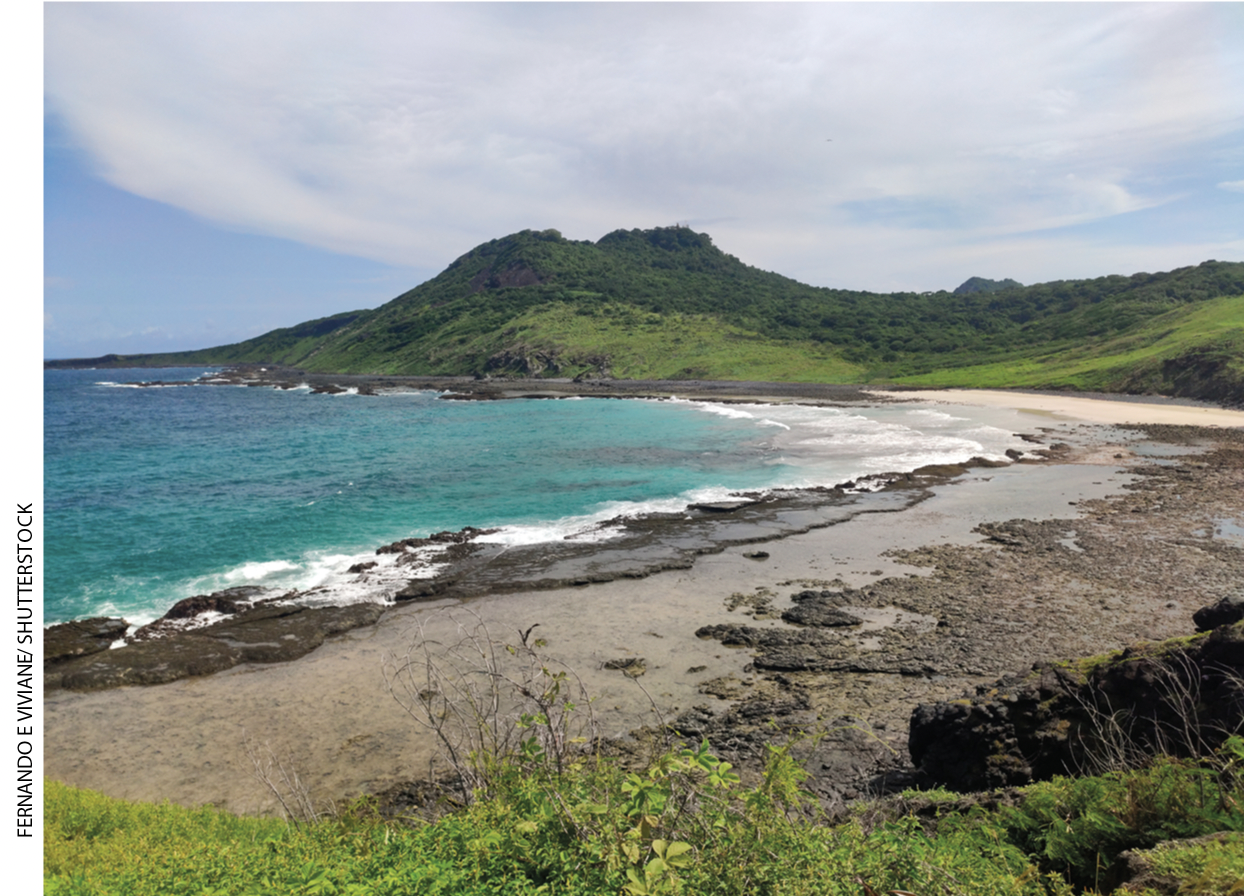 IMAGEM: praia com areia escura, mar azul e, ao fundo, um morro coberto por vegetação. FIM DA IMAGEM.
