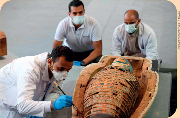 IMAGEM: três profissionais da arqueologia, usando luvas e máscaras, examinam um sarcófago egípcio. um deles trabalha com um pincel sobre o sarcófago. FIM DA IMAGEM.
