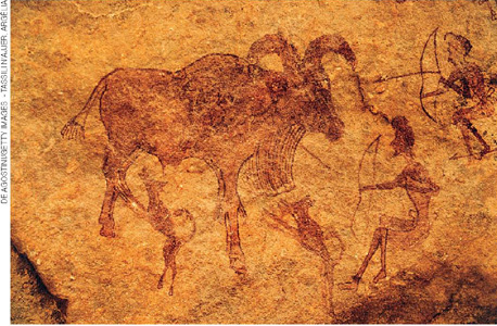 IMAGEM: pintura em rocha, representando dois homens, que apontam seus arcos e flechas em direção a um animal grande como um bovino. animais menores, como cachorros, cercam o bovino. FIM DA IMAGEM.
