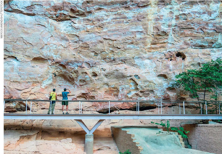 IMAGEM: dois turistas sobre uma ponte de metal em frente a um paredão de pedra o observam e fotografam. FIM DA IMAGEM.
