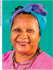 IMAGEM: uma mulher negra, usando brincos, colares e lenço no cabelo, sorri para uma foto. FIM DA IMAGEM.