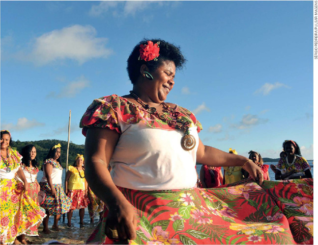 IMAGEM: em primeiro plano, uma mulher negra com flor no cabelo e saia florida está dançando. ao fundo e ao redor dela, outras mulheres participam da dança, usando vestimenta semelhante. FIM DA IMAGEM.