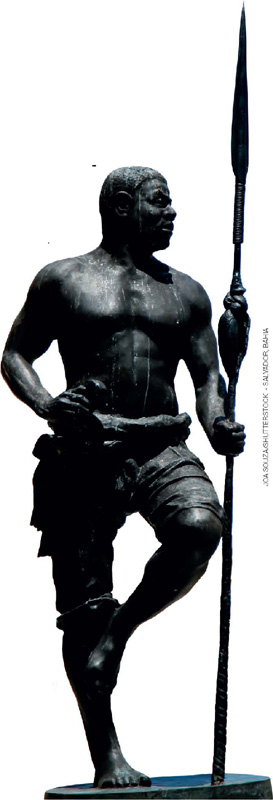 IMAGEM: uma escultura muito alta representando um homem negro e forte. ele está em pé e olha para o lado, com uma das mãos à cintura. com a outra mão, segura uma lança ao lado do corpo. FIM DA IMAGEM.