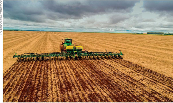 IMAGEM: uma máquina agrícola conduz um arado muito largo, fazendo sulcos no solo em uma propriedade rural. FIM DA IMAGEM.