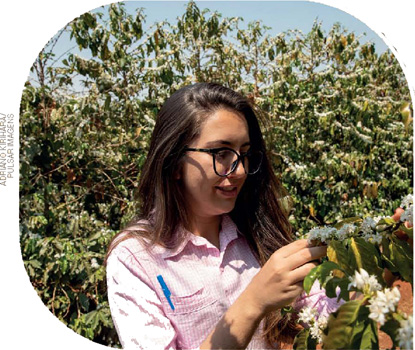IMAGEM: uma mulher de óculos está em um cafezal e examina o galho de uma planta florada. FIM DA IMAGEM.