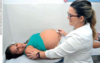 IMAGEM: uma médica examina com um estetoscópio a barriga de uma gestante deitada sobre uma maca. FIM DA IMAGEM.