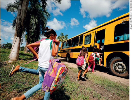 IMAGEM: um ônibus está parado em frente a um terreno gramado, com algumas árvores. crianças com mochilas entram no ônibus. FIM DA IMAGEM.