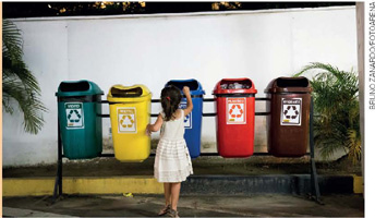 IMAGEM: uma menina joga lixo em uma de cinco lixeiras de coleta seletiva penduradas em um suporte de metal instalado no chão. FIM DA IMAGEM.