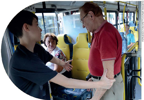 IMAGEM: um garoto oferece o assento em um ônibus a um homem idoso e o apoia pelo braço. no assento ao lado, está sentada uma mulher idosa. FIM DA IMAGEM.