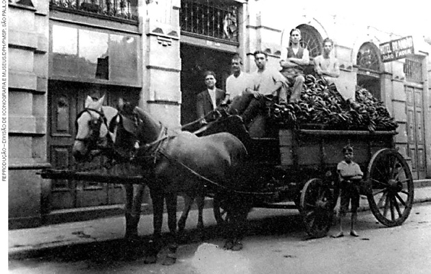 IMAGEM: uma fotografia antiga mostra cinco pessoas em uma carroça, carregada com produtos para venda e puxada por dois cavalos em uma via pública. uma criança está ao lado da carroça. FIM DA IMAGEM.