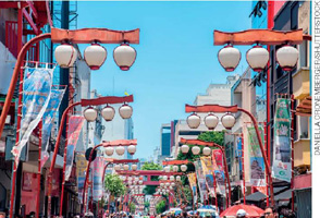 IMAGEM: uma rua com comércio movimentado no bairro da liberdade, na cidade de são paulo. os postes de iluminação pública reproduzem luminárias japonesas, formadas por três cúpulas alinhadas. ao longo da rua, banners com elementos da cultura japonesa. FIM DA IMAGEM.