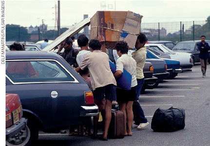 IMAGEM: uma fotografia mostra um estacionamento, onde homens erguem um grande embrulho de papelão para acondicioná-lo no porta-malas de um carro. há bagagem no chão. FIM DA IMAGEM.