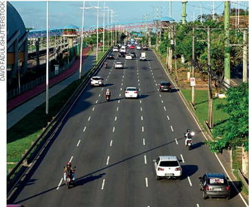 IMAGEM: automóveis trafegam nas cinco faixas de uma pista asfaltada. nas laterais, postes de iluminação pública, ciclovia e árvores. FIM DA IMAGEM.