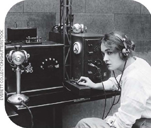 IMAGEM: fotografia em preto e branco do início do século 20. uma mulher com fones de um telégrafo ouve e transmite mensagens. FIM DA IMAGEM.