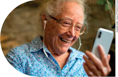 IMAGEM: uma mulher idosa, de óculos e cabelos grisalhos presos, usando fones de ouvido. ela olha para a tela de um celular e sorri. FIM DA IMAGEM.
