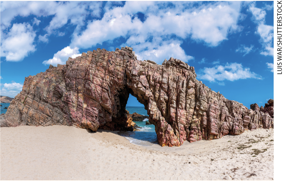 IMAGEM: item b. na areia de uma praia, uma rocha com uma fenda pontiaguda em cima e mais larga embaixo, formando um arco por onde passa água do mar. FIM DA IMAGEM.