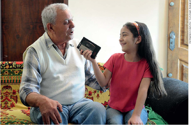 IMAGEM: um homem idoso conta algo a uma menina, que registra a fala do homem com um gravador. FIM DA IMAGEM.