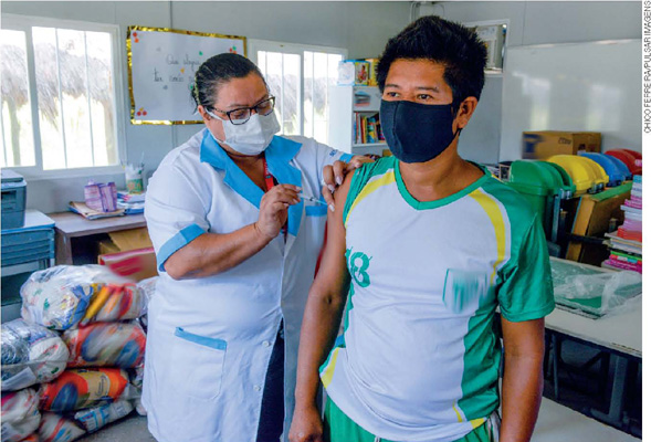 IMAGEM: um homem indígena usando máscara de proteção respiratória é vacinado por uma profissional da saúde uniformizada e também usando máscara. FIM DA IMAGEM.