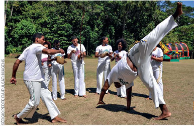 IMAGEM: grupo de jovens joga capoeira em um gramado. alguns deles tocam pandeiro e berimbau. FIM DA IMAGEM.