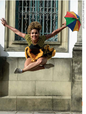 IMAGEM: uma jovem dançarina de frevo salta com as pernas cruzadas e sorri, abrindo os braços e segurando um pequeno guarda-chuva colorido. FIM DA IMAGEM.