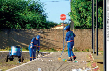 IMAGEM: duas pessoas uniformizadas limpam uma via pública. uma delas varre a calçada. a outra, próxima a um carrinho coletor, recolhe lixo do meio-fio. FIM DA IMAGEM.