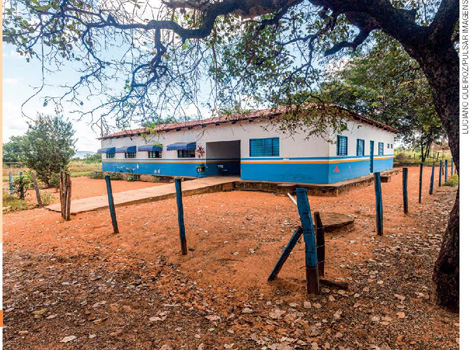 IMAGEM: escola construída sobre uma plataforma baixa de madeira, em um terreno plano e de terra batida, com árvores ao redor. uma rampa dá acesso à entrada da escola, que tem ao seu redor uma cerca de madeira e arame. FIM DA IMAGEM.