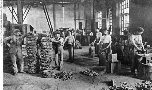 IMAGEM: uma foto em preto e branco mostra um local de produção de ferraduras. à direita, ferreiros usando avental comprido trabalham com martelos em bigornas. à esquerda, dois homens de chapéu empilham materiais. há pequenos amontoados de ferraduras estão no chão. FIM DA IMAGEM.