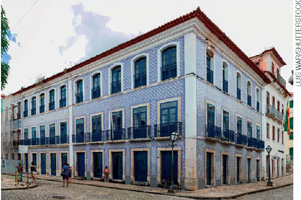 IMAGEM: localizado em uma esquina, um casarão de dois andares com fachada azulejada e janelas amplas e avarandadas. FIM DA IMAGEM.