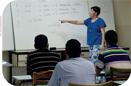 IMAGEM: em uma sala de aula, uma professora explica a matéria escrita em um quadro branco. três alunos adultos prestam atenção nas explicações. FIM DA IMAGEM.