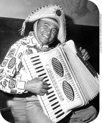 IMAGEM: fotografia em preto e branco. o cantor e compositor luiz gonzaga sorri para a foto enquanto toca sanfona. ele usa um chapéu de couro com uma estrela bordada na aba e veste camisa estampada de mangas compridas. FIM DA IMAGEM.