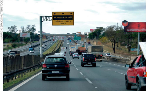 IMAGEM: trânsito intenso de ida e volta em uma rodovia ampla. em uma placa está escrito: amarelo piscante. tráfego lento à frente. FIM DA IMAGEM.