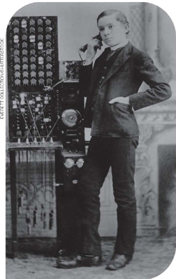 IMAGEM: foto em preto e branco. um homem jovem, de terno e gravata, está em pé, com uma das mãos no bolso. ao lado dele, um aparelho grande, com campainha, botões e um fone, que ele encosta ao ouvido. FIM DA IMAGEM.