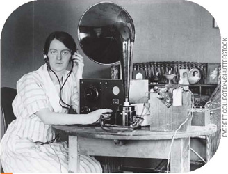 IMAGEM: fotografia em preto e branco do início do século 20. uma mulher está sentada a uma mesa e usa fones de ouvido. ela mexe nos botões de um aparelho cujo som sai por um tubo com o formato de um grande funil dobrado. FIM DA IMAGEM.