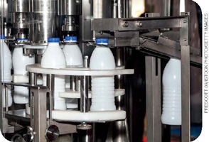 IMAGEM: 1. máquina industrial de envasamento de leite, por onde passam garrafas plásticas em sequência. FIM DA IMAGEM.