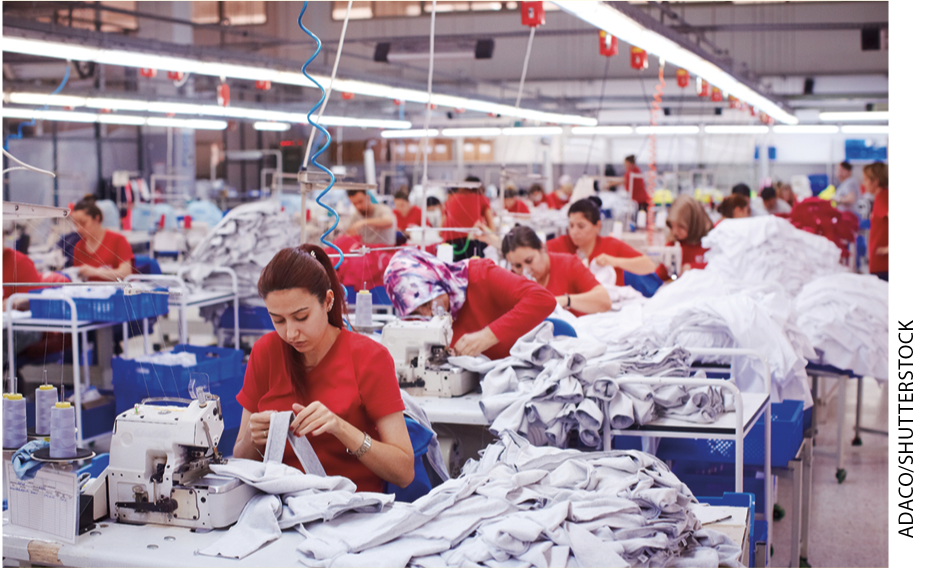 IMAGEM: mulheres uniformizadas trabalham com máquinas de costura na linha de produção de uma fábrica. FIM DA IMAGEM.
