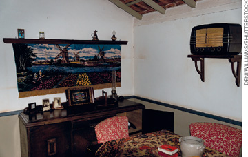 IMAGEM: uma sala com móveis antigos, entre eles: uma mesa com cadeiras, uma cômoda, um rádio e uma obra de arte que retrata uma paisagem holandesa. FIM DA IMAGEM.