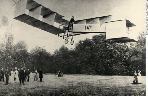 IMAGEM: fotografia antiga de um avião aeroplano em pleno vôo, em um campo aberto. ele é pilotado por um homem, e diversas pessoas assistem. FIM DA IMAGEM.