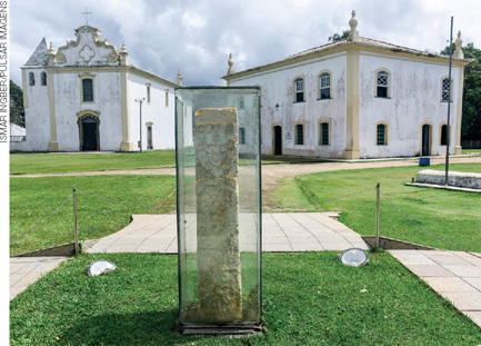 IMAGEM: pequeno monumento de pedra cercado por uma estrutura de vidro em um espaço gramado. ao fundo, há uma igreja. . FIM DA IMAGEM.