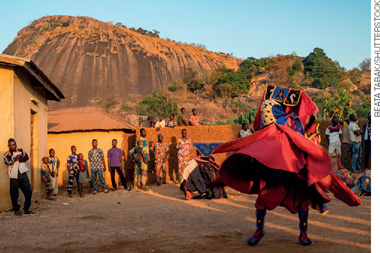 IMAGEM: pessoas em um ambiente aberto em uma celebração, alguns estão ao fundo em pé assistindo um homem dançar sozinho, ele veste roupas com muitos tecidos coloridos e um chapéu caraterístico da cultura africana. FIM DA IMAGEM.