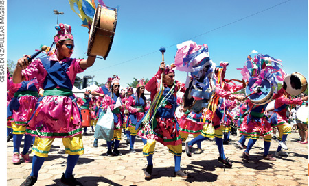 IMAGEM: um grupo de músicos se apresentando em uma festa da congada. eles usam camisas iguais e chapéus com elementos da cultura nordestina, alguns deles seguram tambores com enfeites coloridos. FIM DA IMAGEM.