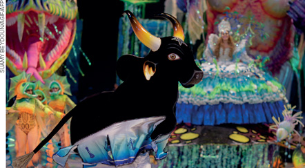 IMAGEM: réplica de um boi se apresentando em um espaço amplo, há pessoas usando fantasias coloridas ao fundo. FIM DA IMAGEM.