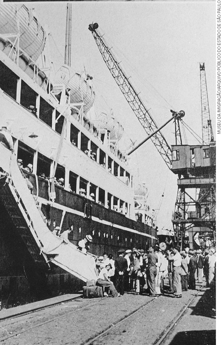 IMAGEM: fotografia antiga de um navio atracado no porto de santos em são paulo, algumas pessoas estão reunidas próximo de uma escada que leva até a embarcação. FIM DA IMAGEM.